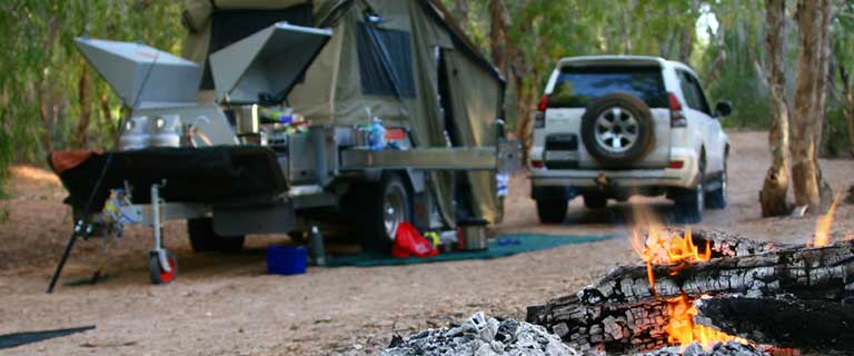 Campfire outside in the woods next to parked car and tent with grill.